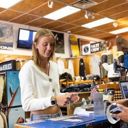 A woman with blonde hair and a white sweater smiles while purchasing an item at a small business. A small business owner in a black baseball cap rings her up.