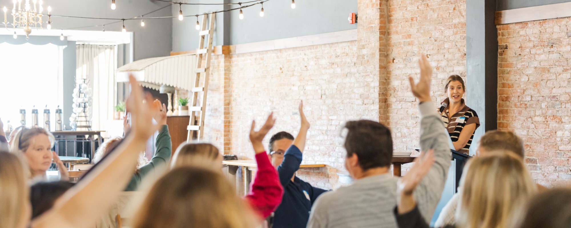 People sitting around tables raise their hands; in the background a speaker holds a microphone.