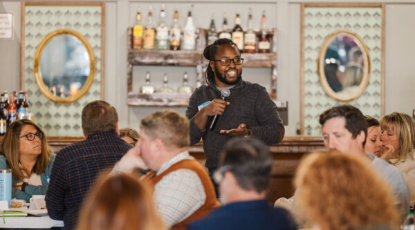 A man stands while speaking into a microphone; he is surrounded by people sitting and listening intently.