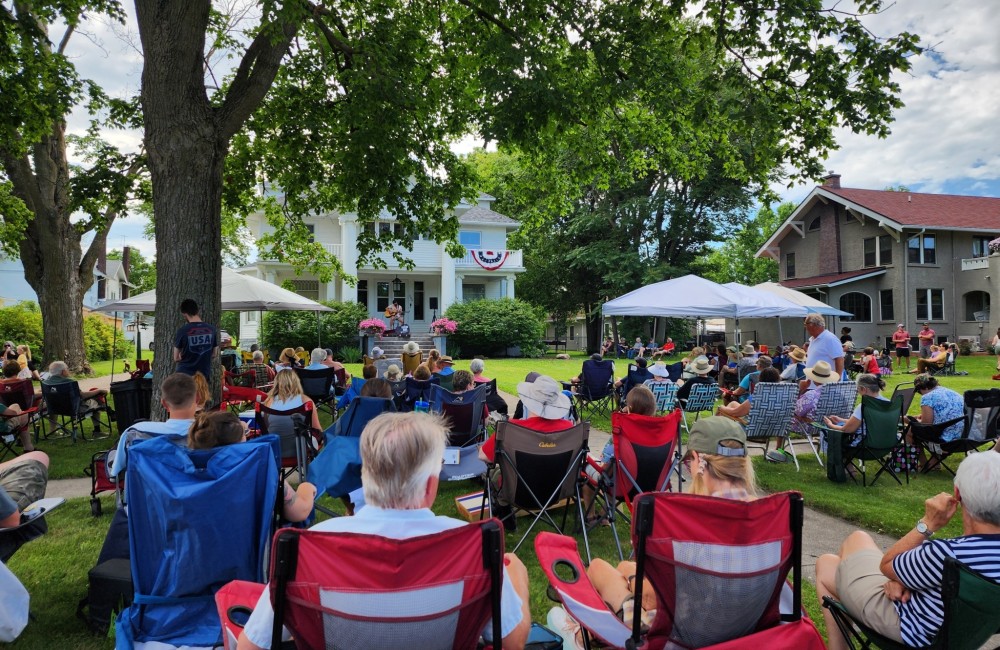 A large group of people are seated in lawn chairs listening to musicians performing from the porch of a home in Jefferson, Iowa.