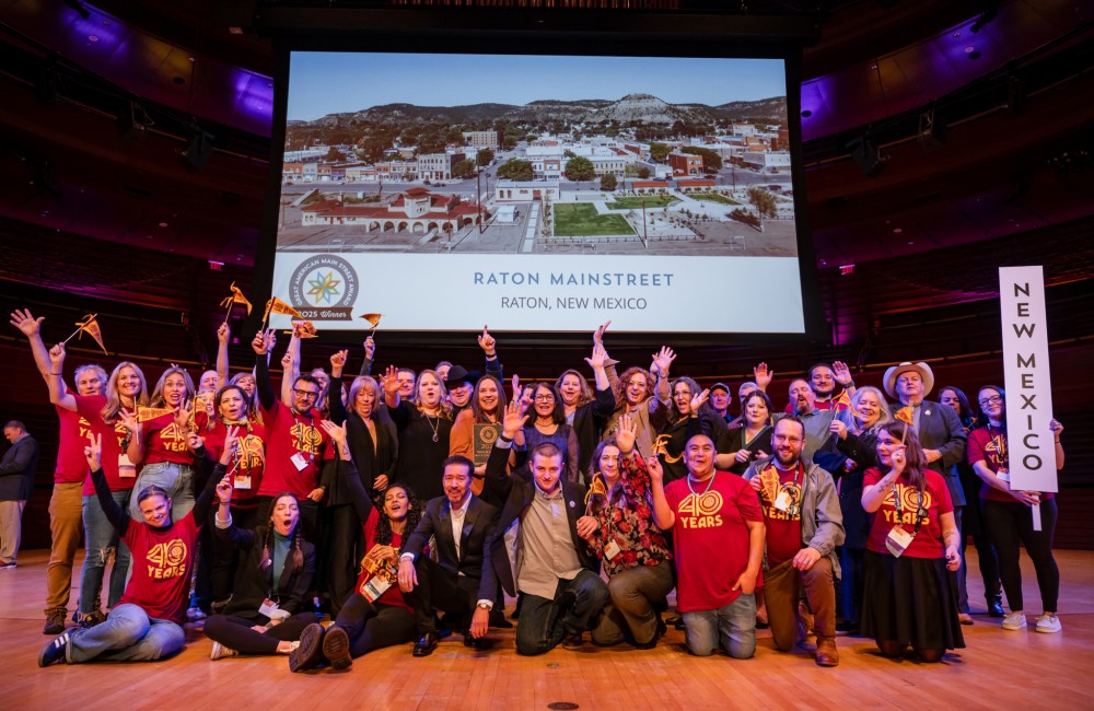 A large group of people stand on a stage, waving flags and cheering for Raton Mainstreet's 2025 Great American Main Street Award.