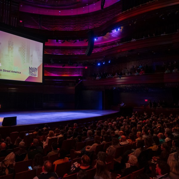 A large crowd sitting in a dimly-lit theater listening to a person speaking from the stage.