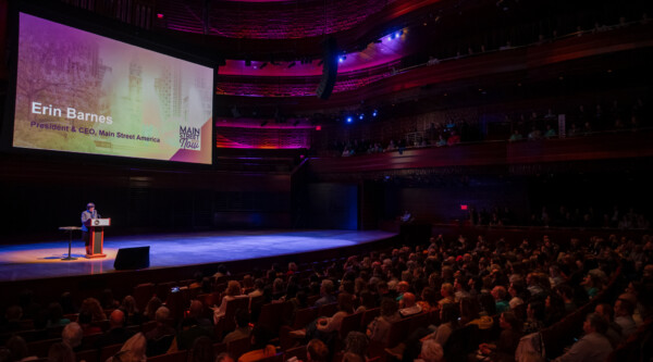 A large crowd sitting in a dimly-lit theater listening to a person speaking from the stage.