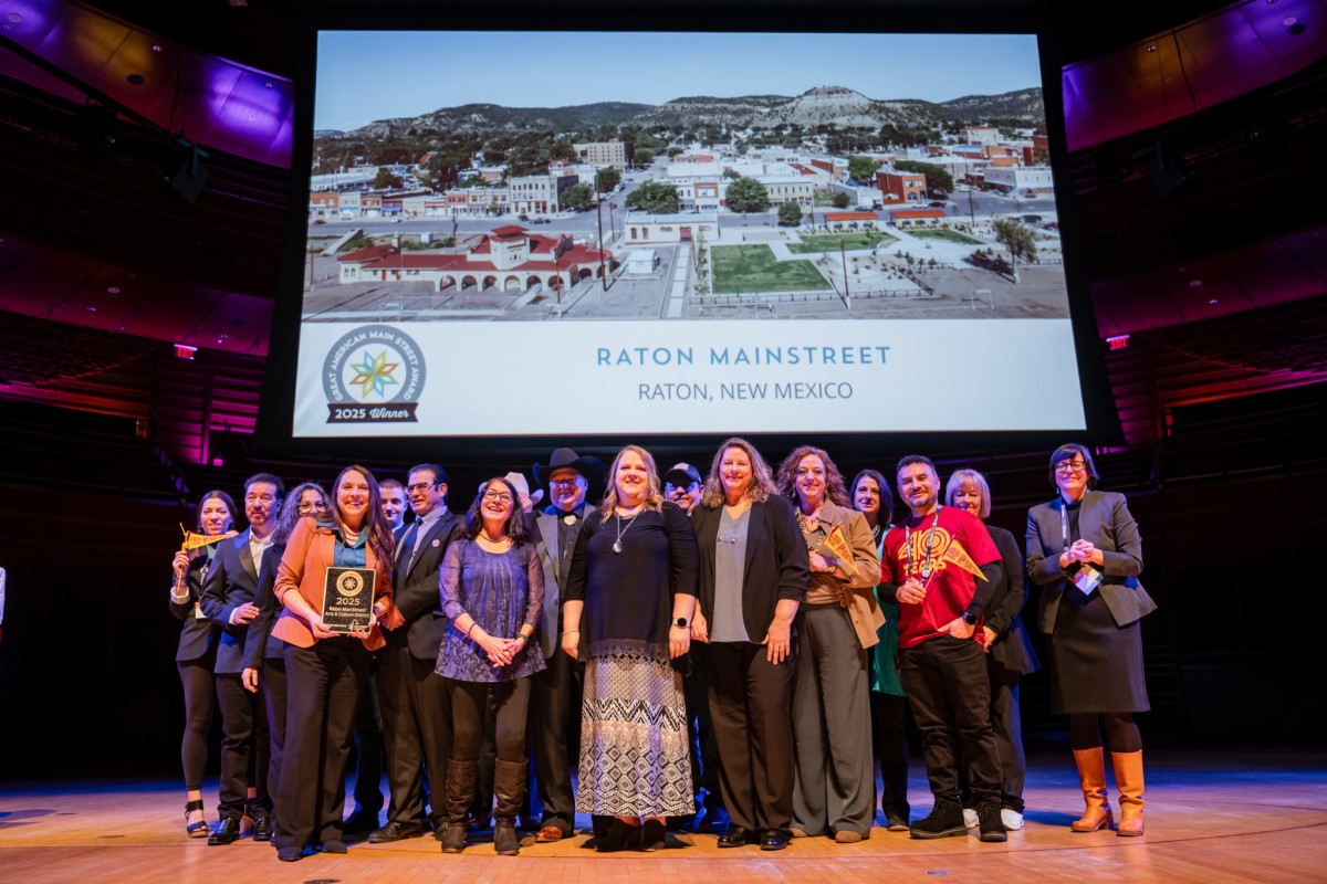 Large group on stage celebrating award. Large screen behind group reads "RATON MAINSTREET - RATON, NEW MEXICO."