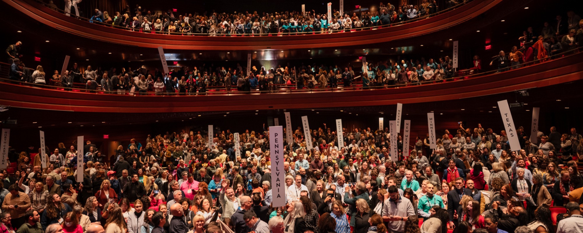 A large theater filled with people, some holding up signs displaying the names of US states.