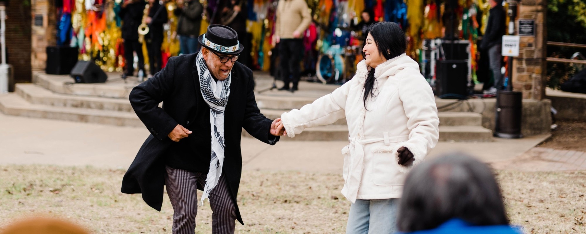 At an outdoor concert event, a man and woman dance in front of a small amphitheater stage decorated with colorful streamers where a band is performing.