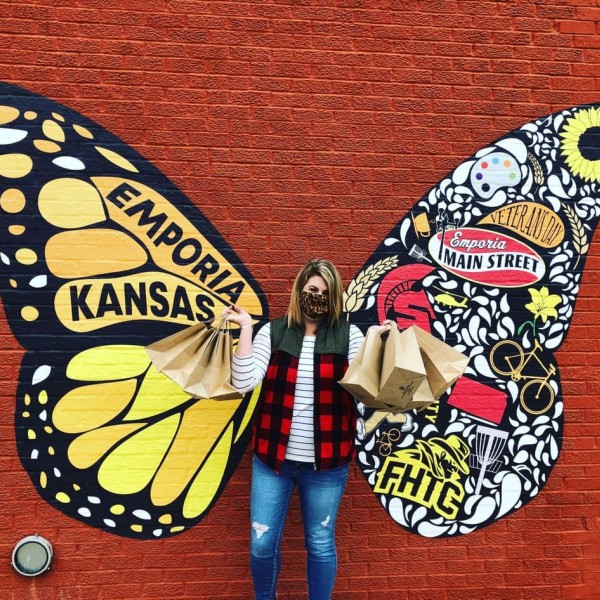 A woman wearing a mask and holding paper gift bags stands in front of a large butterfly mural.