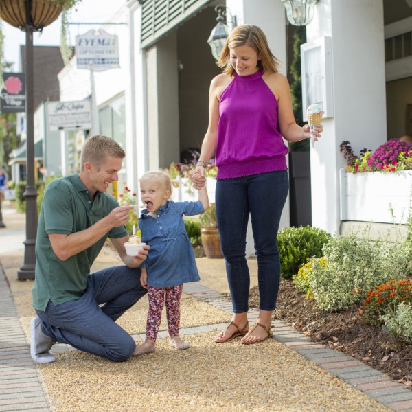 A father offers a spoonful of ice cream to his young daughter while she holds her mother's hand.