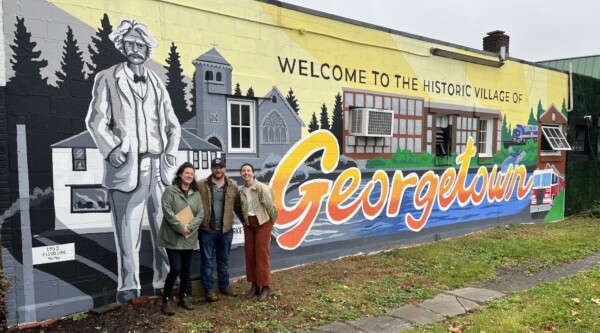 Three people posing in front of a large mural with scenes of Georgetown Connecticut