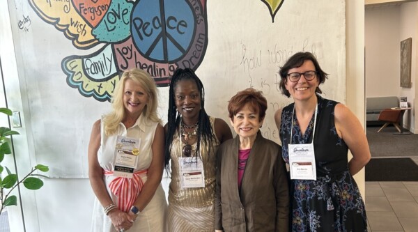 Four women stand in front of a mural depicting a peace dove.