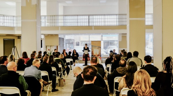 A woman stands at a podium while giving a presentation to dozens of people sitting in chairs theater style.