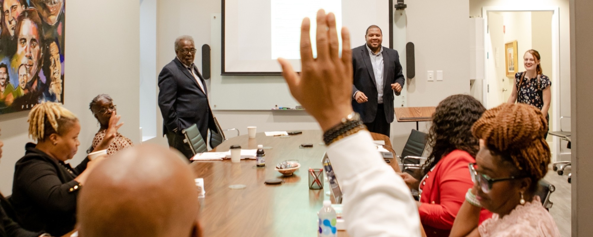 People sit around a conference table; a participant raises their hand while presenters speak from the front of the room.