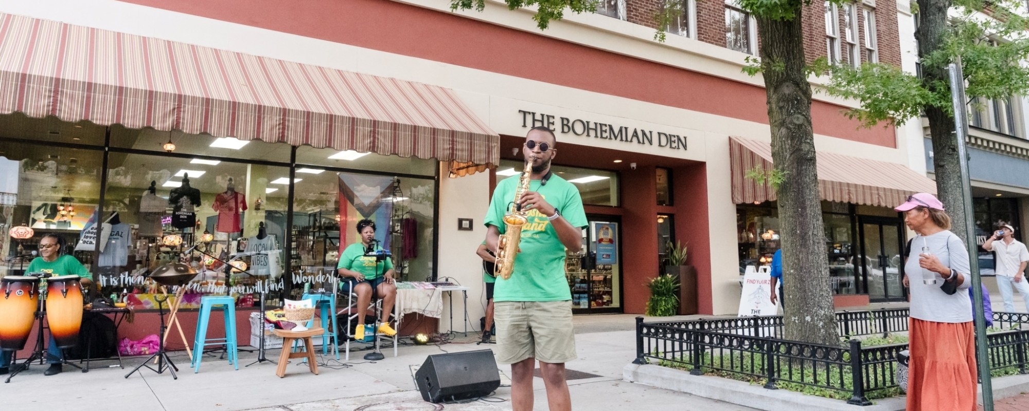 A saxophonist, drummer, and vocalist set-up on a wide sidewalk perform for pedestrians.