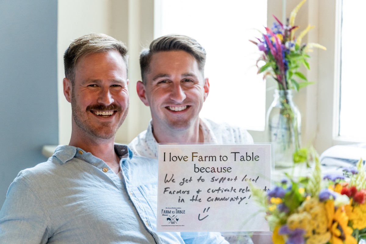 Two men sit side-by-side and smile while holding up a sign with a handwritten comment.