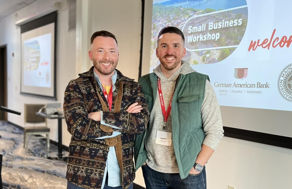 Two men smile while stand side-by-side and in front of a projection displaying the name of a workshop.