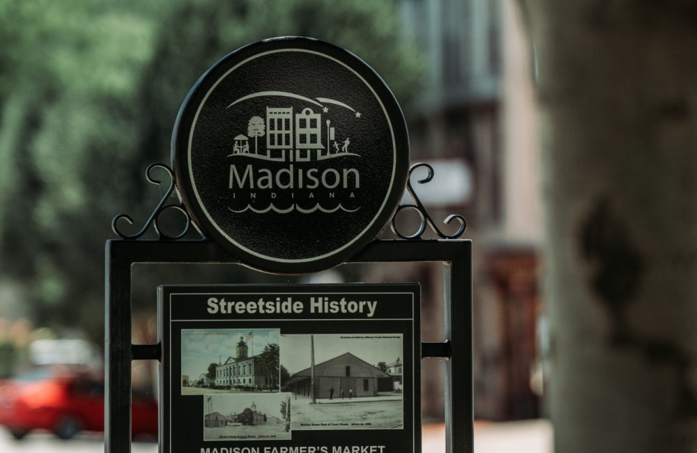 Metal historic sidewalk marker next to a tree.