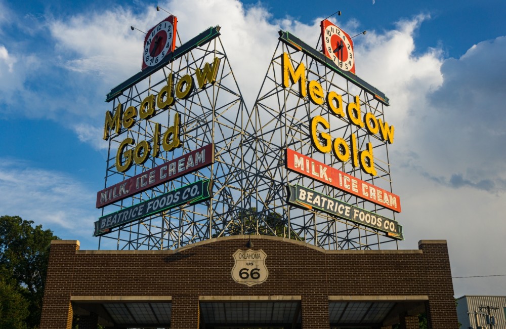 Colorful, two-sided metal structure sign for Meadow Gold products located on top of a brick building with a Route 66 emblem.