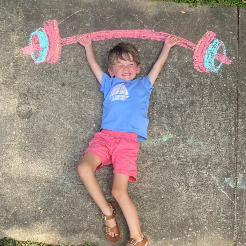 A young boy lays on a sidewalk with his arms raised above his head to hold up a chalk drawing of a dumbbell.