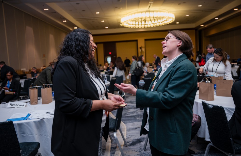 Two women chat while standing in a large seminar room filled with people.