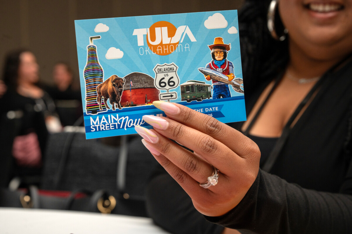 A woman holding up a "save the date" postcard for the 2026 Main Street Now Conference.