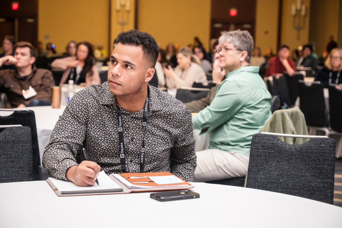 People sit around tables while listening to a conference speaker; in the foreground, a man looks up while taking notes.