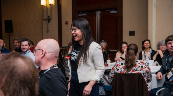 A woman smiles while walking between tables filled with people intently listening to a presenter.