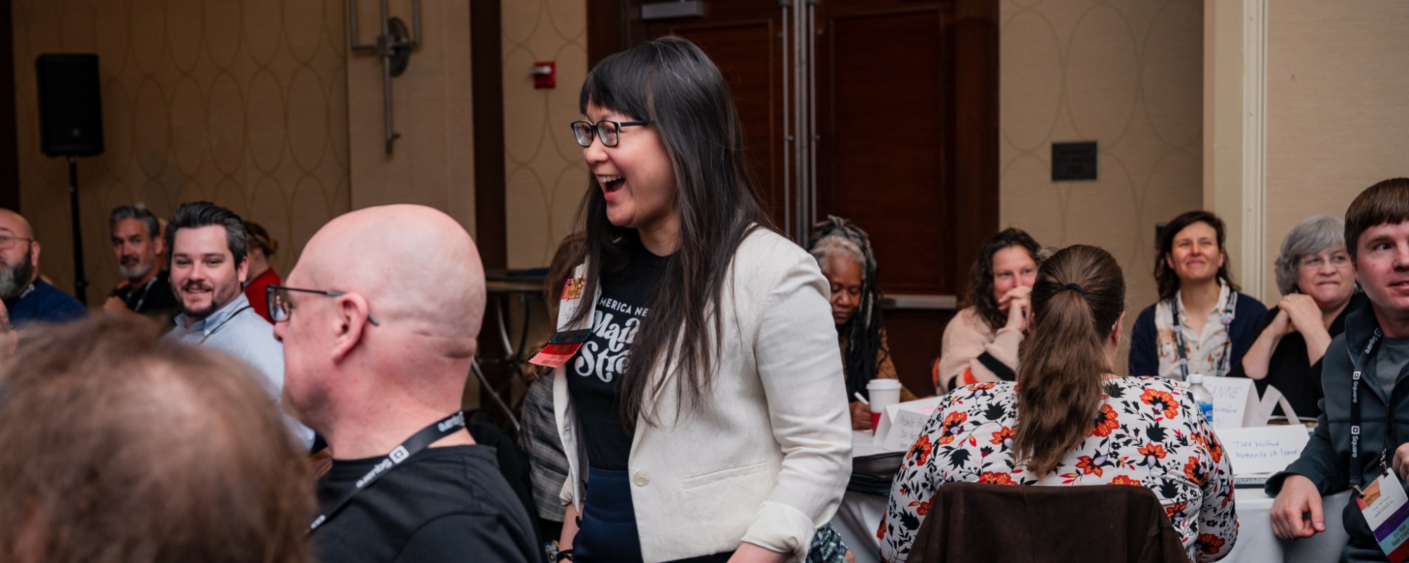 A woman smiles while walking between tables filled with people intently listening to a presenter.