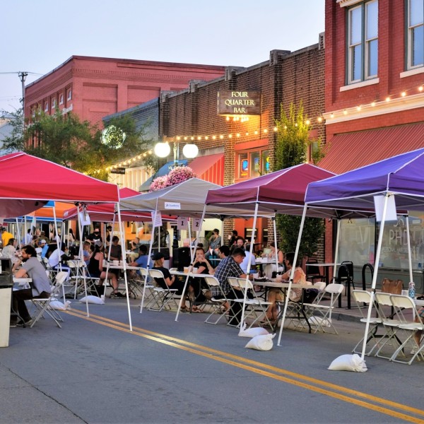 People gather around tables set-up underneath brightly colored pop-up canopy tents that have been arranged in a single line down the middle of a downtown street.