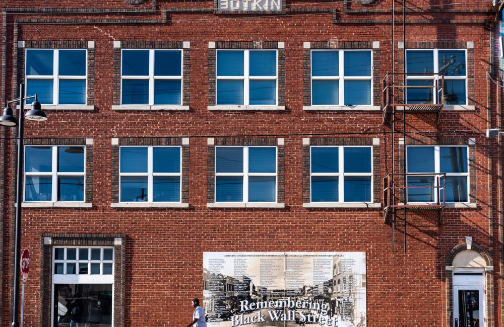 A person walks past a large brick building with a mural featuring a black-and-white photo of Tulsa's historic Black Wall Street district and the words "Remembering Black Wall Street" in Gothic-style font.