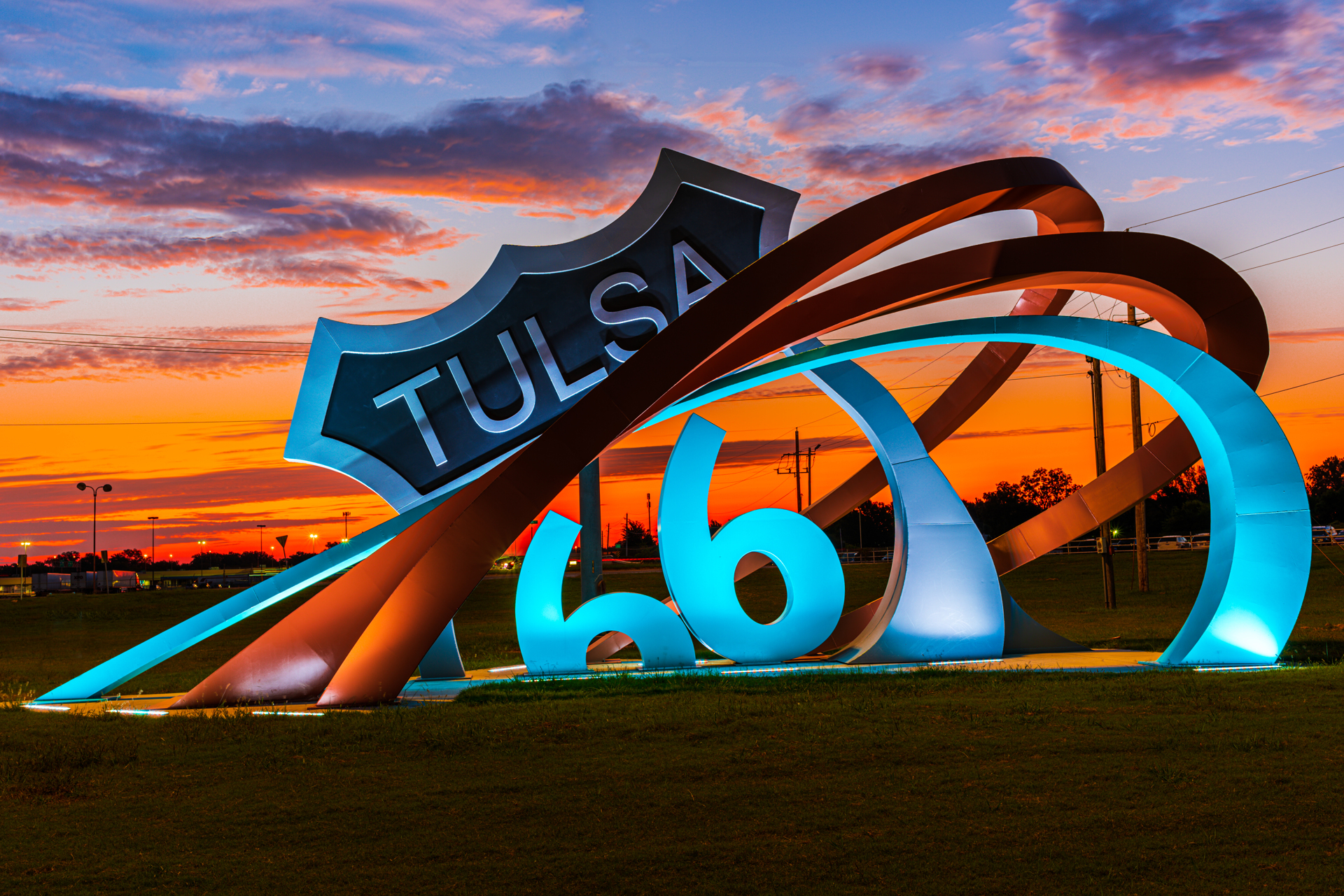 Large sculpture consisting of three metal bands rising from the ground (left) and curving around a replica of a "Tulsa Route 66" sign (center). The sculpture is located in an open grassy area and is illuminated by ground lights.