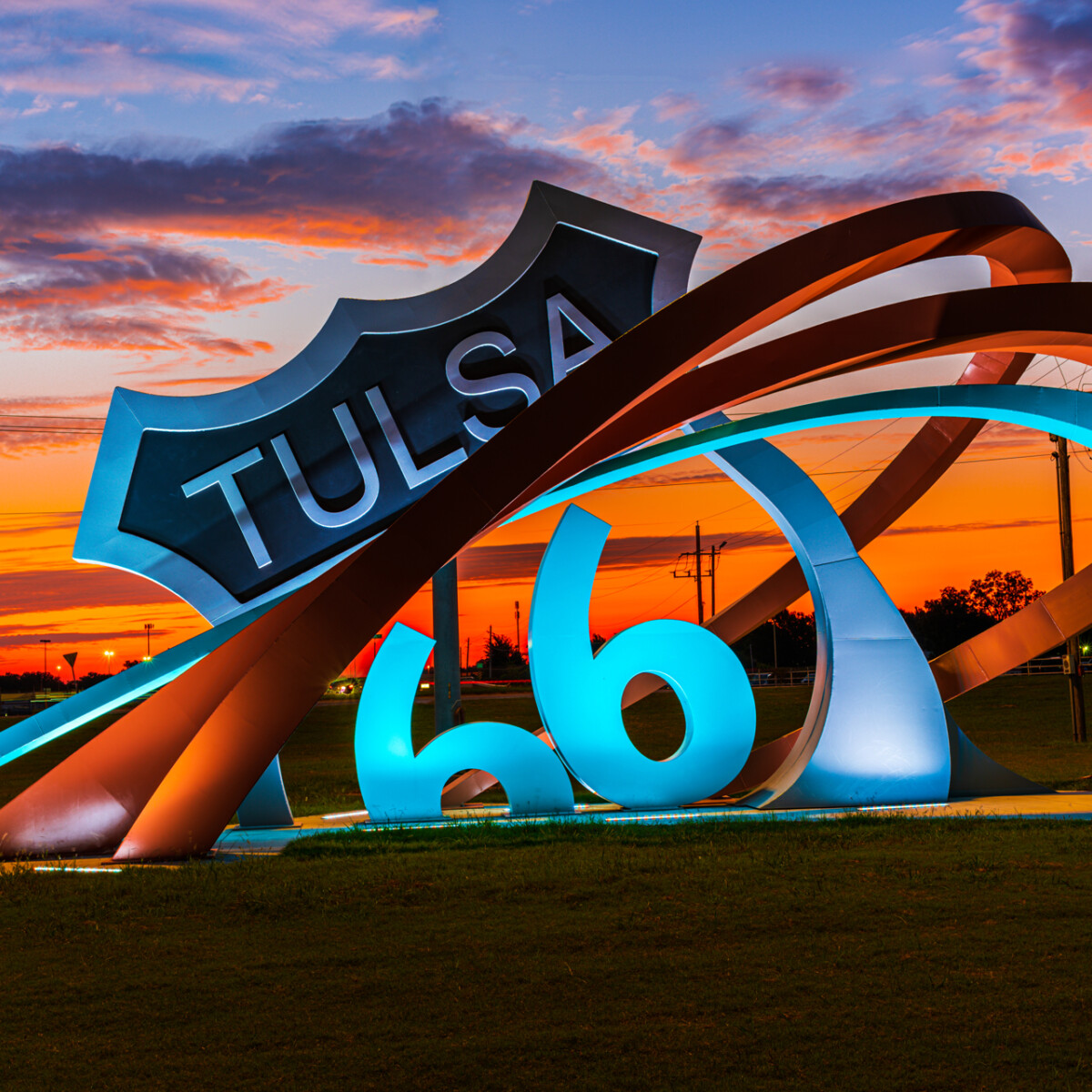 Large sculpture consisting of three metal bands rising from the ground (left) and curving around a replica of a "Tulsa Route 66" sign (center). The sculpture is located in an open grassy area and is illuminated by ground lights.