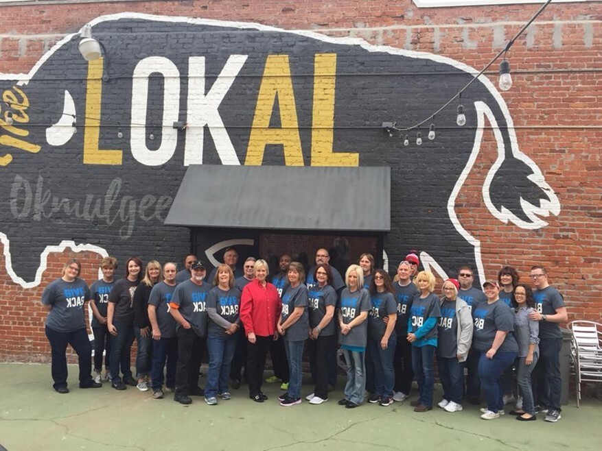 A large group of people posing in front of a mural of a buffalo with the words "lOKal okmulgee"