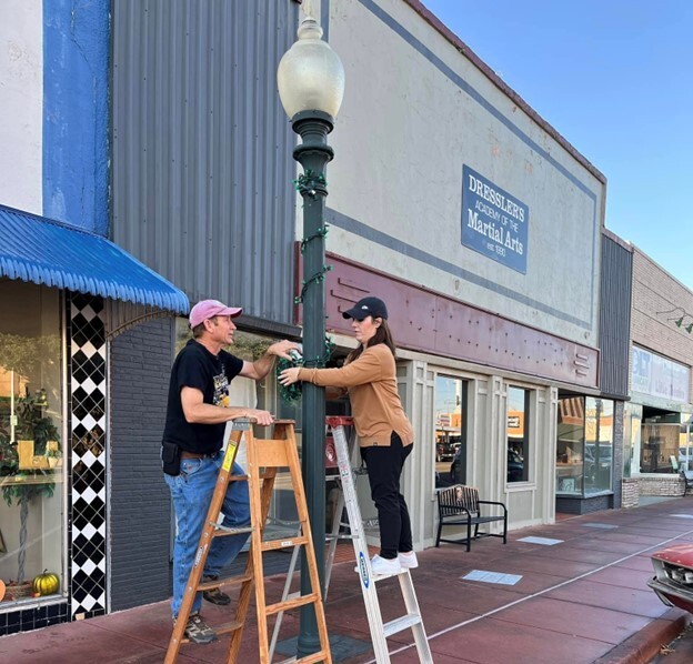 Two people on ladders work together to put Christmas lights on a downtown lamp post