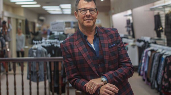 A man wearing glasses and a plaid suit jacket leans on a flight of stairs and smiles, standing in a clothing shop.