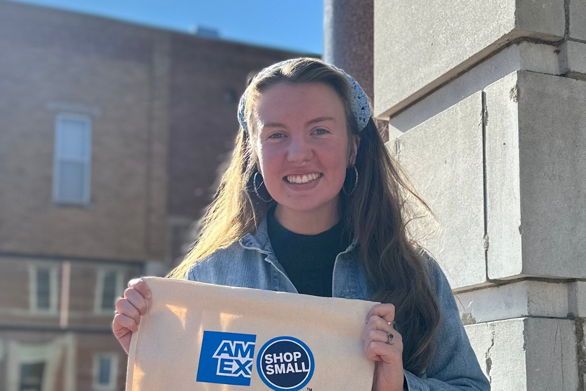Smiling woman holds bag with "AmEx Shop Small" branding in front of storefront.