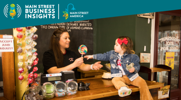 Woman behind counter at candy store gives lollipop to child sitting on counter.